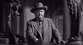 Movie still from “3:10 to Yuma” (1957), directed by Delmer Daves – An older man wearing a suit and a hat holding a glass; Medium shot, Low angle