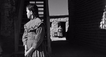 Movie still from “3:10 to Yuma” (1957), directed by Delmer Daves – A woman standing in front of a brick building; Wide shot, Over the shoulder angle