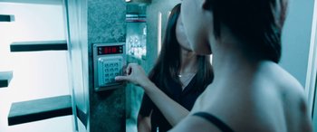 Movie still from “4.3.2.1.” (2010), directed by Mark Davis – Two women are standing in a room and one of them is pressing a button on the wall; Close Up shot, Over the shoulder angle