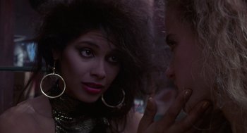 Movie still from “52 Pick-Up” (1986), directed by John Frankenheimer – A woman with big earrings and a woman with very long hair looking at each other in a dark room; Close Up shot, Over the shoulder angle