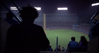 Movie still from “52 Pick-Up” (1986), directed by John Frankenheimer – A man standing in a stadium watching a baseball game at night; Wide shot, Over the shoulder angle