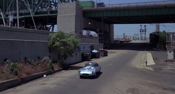 Movie still from “52 Pick-Up” (1986), directed by John Frankenheimer – A car driving down a street next to a bridge; Extreme Wide shot, High angle