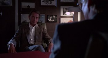 Movie still from “52 Pick-Up” (1986), directed by John Frankenheimer – A man sitting in front of another man in front of a wall; Medium shot, Over the shoulder angle