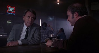 Movie still from “52 Pick-Up” (1986), directed by John Frankenheimer – Two men sitting at a table in front of a projector screen; Medium shot, Over the shoulder angle