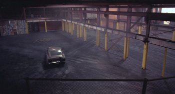 Movie still from “52 Pick-Up” (1986), directed by John Frankenheimer – A car parked in a parking lot next to a metal fence; Extreme Wide shot, High angle