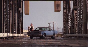 Movie still from “52 Pick-Up” (1986), directed by John Frankenheimer – A man sitting on the hood of a blue car; Wide shot, Low angle