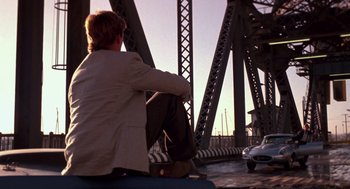 Movie still from “52 Pick-Up” (1986), directed by John Frankenheimer – A man sitting on the side of a bridge; Wide shot, Over the shoulder angle