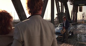 Movie still from “52 Pick-Up” (1986), directed by John Frankenheimer – A man standing on a bridge looking at a car; Wide shot, Over the shoulder angle