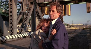 Movie still from “52 Pick-Up” (1986), directed by John Frankenheimer – A man and a woman hugging in front of a bridge; Medium shot, Low angle