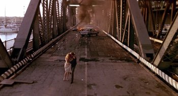Movie still from “52 Pick-Up” (1986), directed by John Frankenheimer – A couple kissing on a bridge with a car on fire in the background; Wide shot, High angle