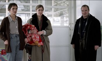 Movie still from “71 Fragments of a Chronology of Chance” (1994), directed by Michael Haneke – A woman in a trench coat holding a baby; Medium shot, Over the shoulder angle