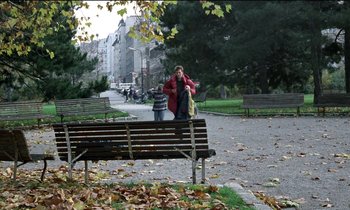 Movie still from “71 Fragments of a Chronology of Chance” (1994), directed by Michael Haneke – A woman standing next to a bench on a sidewalk; Extreme Wide shot, High angle