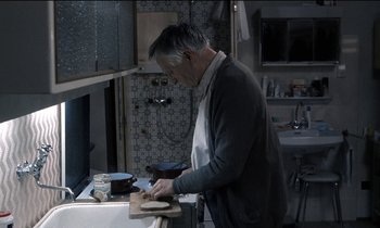 Movie still from “71 Fragments of a Chronology of Chance” (1994), directed by Michael Haneke – An older man in a kitchen preparing a meal; Medium shot, High angle