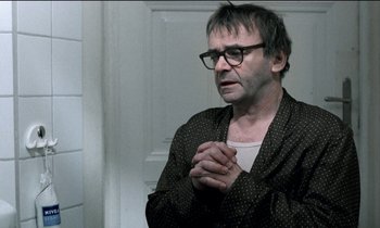 Movie still from “71 Fragments of a Chronology of Chance” (1994), directed by Michael Haneke – A man with glasses is praying in front of a door; Medium shot, Over the shoulder angle