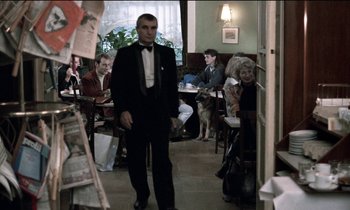 Movie still from “71 Fragments of a Chronology of Chance” (1994), directed by Michael Haneke – A man in a tuxedo standing next to a table; Wide shot, Over the shoulder angle