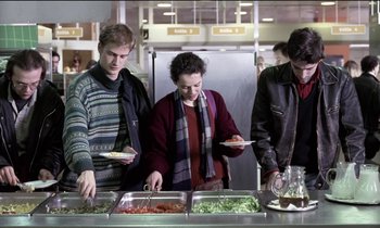 Movie still from “71 Fragments of a Chronology of Chance” (1994), directed by Michael Haneke – A group of people standing in front of a buffet line; Medium shot, Over the shoulder angle