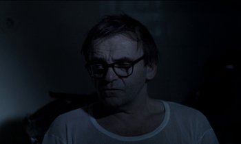 Movie still from “71 Fragments of a Chronology of Chance” (1994), directed by Michael Haneke – A man wearing glasses and a white t - shirt; Close Up shot, Over the shoulder angle