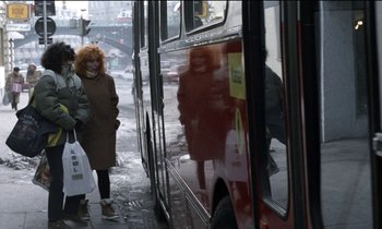 Movie still from “71 Fragments of a Chronology of Chance” (1994), directed by Michael Haneke – A woman walking on the sidewalk next to a red bus; Wide shot, Over the shoulder angle