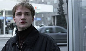 Movie still from “71 Fragments of a Chronology of Chance” (1994), directed by Michael Haneke – A young man standing in front of a building; Close Up shot, Low angle