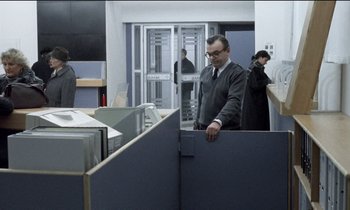 Movie still from “71 Fragments of a Chronology of Chance” (1994), directed by Michael Haneke – A man standing in an office cubicle looking at the floor; Wide shot, Over the shoulder angle