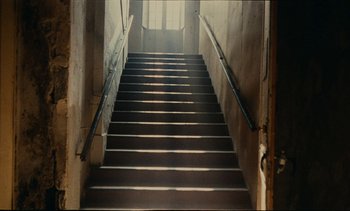 Movie still from “7 Rm., kit., ba...” (1984), directed by Agnès Varda – A staircase with a railing going up the side of it; Wide shot, Low angle