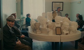 Movie still from “7 Rm., kit., ba...” (1984), directed by Agnès Varda – A group of people sitting around a table with many heads on it; Wide shot, High angle