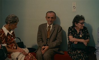 Movie still from “7 Rm., kit., ba...” (1984), directed by Agnès Varda – An older man sitting next to two older women; Wide shot, High angle