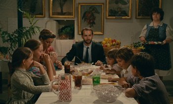 Movie still from “7 Rm., kit., ba...” (1984), directed by Agnès Varda – A group of people sitting around a table eating; Medium shot, Over the shoulder angle