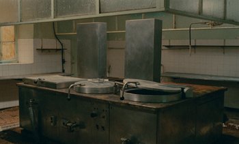 Movie still from “7 Rm., kit., ba...” (1984), directed by Agnès Varda – Two sinks in an industrial style kitchen with metal fixtures; Wide shot, High angle