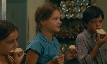 Movie still from “7 Rm., kit., ba...” (1984), directed by Agnès Varda – A young girl holding a piece of food in her hands; Medium shot, Over the shoulder angle