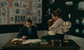 Movie still from “7 Rm., kit., ba...” (1984), directed by Agnès Varda – A man sitting at a desk with a woman and a baby; Medium shot, Over the shoulder angle