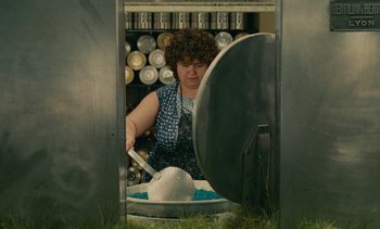 Movie still from “7 Rm., kit., ba...” (1984), directed by Agnès Varda – A woman in a blue apron is cutting a cake; Medium shot, Low angle