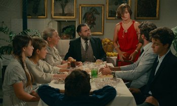 Movie still from “7 Rm., kit., ba...” (1984), directed by Agnès Varda – A group of people sitting around a table with food; Wide shot, High angle