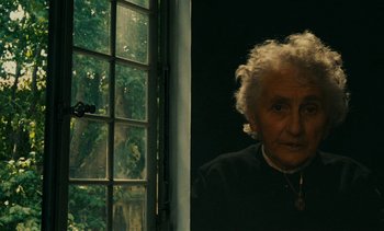 Movie still from “7 Rm., kit., ba...” (1984), directed by Agnès Varda – An older woman standing in front of a window; Close Up shot, Over the shoulder angle