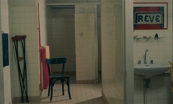Movie still from “7 Rm., kit., ba...” (1984), directed by Agnès Varda – A black chair in a bathroom next to a tiled shower; Extreme Wide shot, High angle