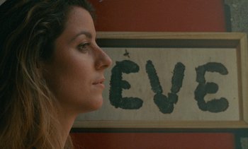 Movie still from “7 Rm., kit., ba...” (1984), directed by Agnès Varda – A woman's face in front of an eve sign; Close Up shot, Low angle