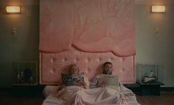 Movie still from “7 Rm., kit., ba...” (1984), directed by Agnès Varda – A man and a woman are sitting in a bed reading magazines; Wide shot, Overhead angle