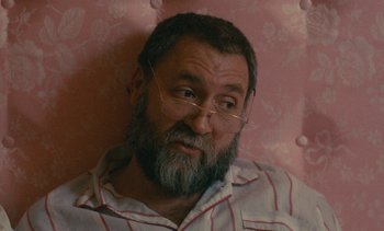 Movie still from “7 Rm., kit., ba...” (1984), directed by Agnès Varda – A man with a beard and glasses is sitting on a chair; Close Up shot, High angle