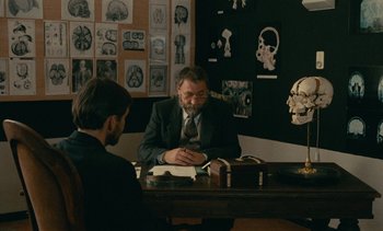Movie still from “7 Rm., kit., ba...” (1984), directed by Agnès Varda – Two men sitting at a table in front of a book; Medium shot, Over the shoulder angle