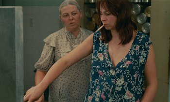 Movie still from “7 Rm., kit., ba...” (1984), directed by Agnès Varda – A woman holding a cigarette in front of another woman; Medium shot, Over the shoulder angle