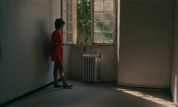 Movie still from “7 Rm., kit., ba...” (1984), directed by Agnès Varda – A woman in a red dress looking out of a window; Wide shot, High angle