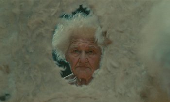 Movie still from “7 Rm., kit., ba...” (1984), directed by Agnès Varda – An old woman with a white wig looking at the camera through a hole in the wall; Close Up shot, Low angle