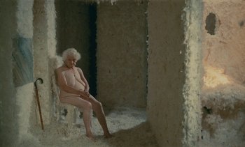 Movie still from “7 Rm., kit., ba...” (1984), directed by Agnès Varda – An old woman sitting naked in a room; Wide shot, High angle