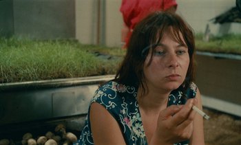 Movie still from “7 Rm., kit., ba...” (1984), directed by Agnès Varda – A woman sitting on the ground looking at her cell phone; Close Up shot, High angle