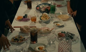 Movie still from “7 Rm., kit., ba...” (1984), directed by Agnès Varda – A table filled with lots of food and glasses of beverages; Medium shot, High angle