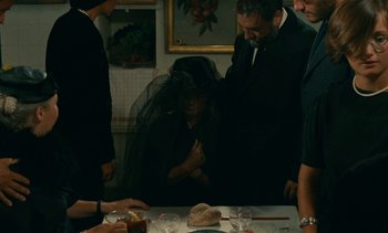 Movie still from “7 Rm., kit., ba...” (1984), directed by Agnès Varda – A group of people standing around a table with food on top of it; Medium shot, Over the shoulder angle