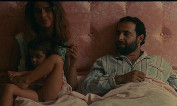 Movie still from “7 Rm., kit., ba...” (1984), directed by Agnès Varda – A man and a woman sitting in bed next to a little girl; Medium shot, High angle