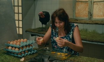 Movie still from “7 Rm., kit., ba...” (1984), directed by Agnès Varda – A woman in a blue dress is peeling an egg; Medium shot, High angle