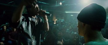 Movie still from “8 Mile” (2002), directed by Curtis Hanson – A crowd of people are gathered around a man singing into a microphone; Medium shot, Over the shoulder angle