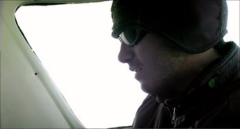 Movie still from “9 Songs” (2004), directed by Michael Winterbottom – A man wearing a hat and sunglasses looking out a window; Close Up shot, High angle